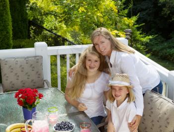 Front view of mature mother holding her two happy daughters while eating breakfast outdoors, during summer time, on patio with woods in background 