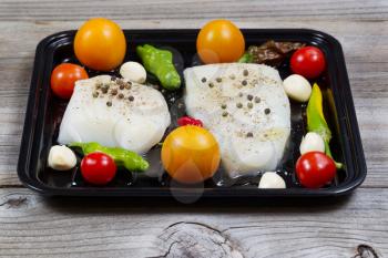 Front view of fresh raw Cod Fish Fillets in cooking pan with tomatoes, garlic, peppers and peppercorn salt on rustic wooden boards 
