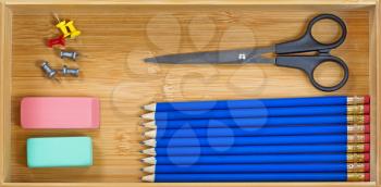 Top view of blue colored pencils, lined up, eraser, scissors, and pin tacks inside wooden desk drawer 