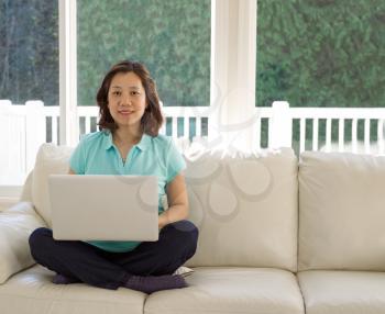 Mature woman, looking forward, sitting on her white couch while using her laptop in her family room with bright sunlight coming through windows and trees blurred out in background.