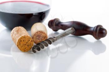 Close up front view of antique corkscrew, focus on front part of corkscrew and cork on far left side, on glass table with red wine in glass in background along with reflections 