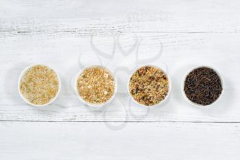Top view of various rice types each within a small bowl on white wood