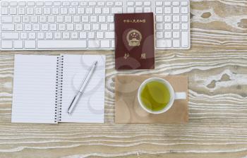 Top view shot of working desktop, China passport, keyboard and green tea drink on aged white wood surface in horizontal format.