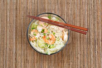 Top view image of Chinese noodle soup in clear glass bowl on top of natural bamboo placemat. Layout in horizontal format.