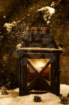 Vintage concept of an old metal lantern, glowing red candle inside, with soft falling snow and an evergreen tree branch on rustic wood in background 