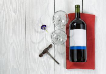 Overhead angled view of a large bottle of red wine, drinking glasses, serving napkin, flower and antique corkscrew on white wooden boards. 