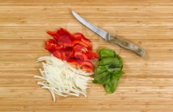 High angled view of freshly sliced red pepper, white onion, and large basil leafs with cutting knife on natural bamboo board. 