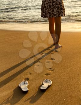 Woman, waist down, walking barefoot into Pacific Ocean during golden sunset. Focus on foot prints with sandals in forefront. 