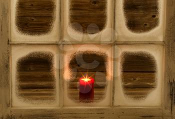 Snow covered window and glowing red candle on rustic wood background. 