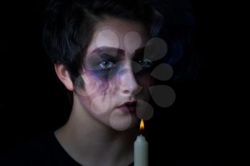 Teen girl in scary makeup holding candle with flame on black background. 