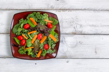 High angled view of fresh green salad with carrots, cherry tomato, basil, baby kale, and lettuce on rustic white wooden boards.  