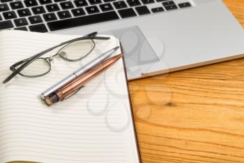 Close up of pens, selective focus on pens, with open blank notebook, reading glasses, and computer in background on desktop. 