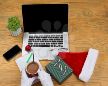 High angled view of hands wearing white gloves while preparing to drink hot chocolate with computer, present, cap, notepad, pencil and plant on desktop. Christmas concept of Santa Claus office. 