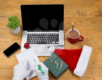High angled view of Santa Claus writing his gift list with hot chocolate, computer, present, cap, notepad, pencil, cell phone, reading glasses and plant on desktop. Christmas concept of Santa Claus of