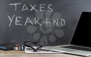 Desktop with tax form, laptop, reading glasses, wrist watch, pens, chalkboard, and calculator. Focus on front part of desk objects. 