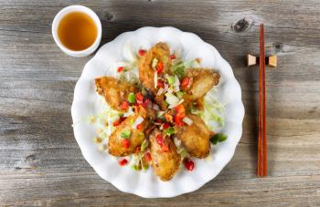 Top view of fried Asian style chicken wings in white plate with garnishes. Green tea and chopsticks in holder. Rustic wooden boards underneath. 