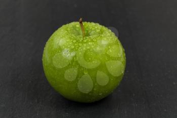 Close up of a fresh whole green apple, covered with water droplets, with natural slate stone underneath. Selective focus on front part of fruit. 