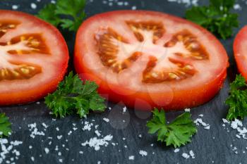 Close up front view of juicy slices of a ripe tomato, parsley and sea salt flakes on natural stone slate. 