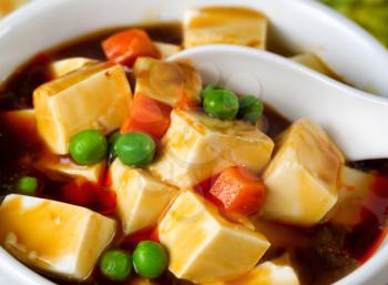 Close up view of tofu, peas, and carrots in soup with spoon in bowl. 