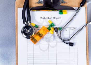 Patient medication record form with stethoscope, pills, container, clipboard on stainless steel counter. 
