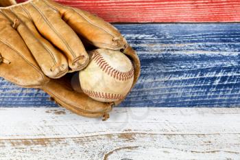 Close up of old worn baseball mitt and baseball on faded wooden boards painted red, white and blue. 