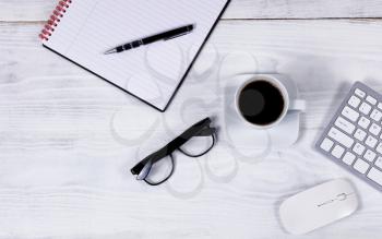 Overhead view of white desktop with partial keyboard, black coffee, reading glasses, notepad, pen and computer mouse. 
