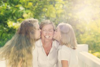Front view of mother receiving kisses from her daughters while outside on outdoor patio. Haze light effect applied to image.


