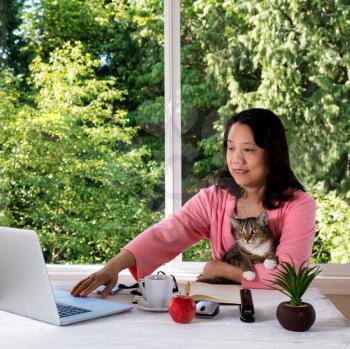 Mature woman, wearing morning attire, holding her family pet cat while working from home in front of large window with bright daylight and trees in background. 