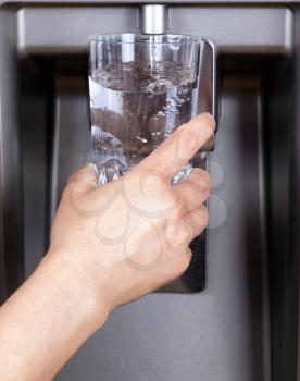 Hand holding drinking glass while being filled with filtered water from refrigerator