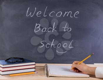 Hand holding pencil with notepad on desktop. Erased black chalkboard in background along with books, pencils, and reading glasses. 