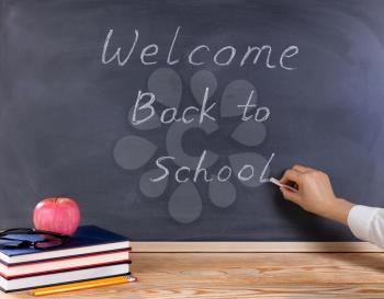 Teacher hand writing on erased black chalkboard. Desktop with books, apple, pencils, and reading glasses in forefront. Back to school concept. 