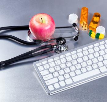 Medical stethoscope on exam table with computer keyboard, medicine pills, bottles and apple.  