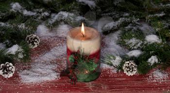 Close up of a burning candle with snowy fir branches and pine cones on rustic red wooden boards for Christmas concept. 
