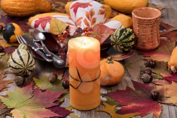 Selective focus of a close up view of holiday candle burning with autumn dinner setting with real gourd decorations, leaves and acorns on top of rustic wood