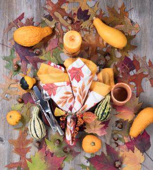 Top view of autumn dinner setting with burning candle, cup, real gourd decorations, leaves and acorns on top of rustic wood