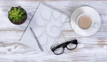 Notebook and pen with reading glasses, plant and creamy coffee on rustic white table