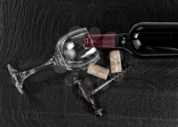 Overhead view of antique wine corkscrew, red wine bottle, glass and used corks on black slate 