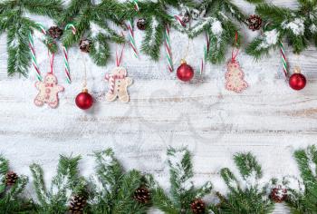 Snowy Christmas evergreen branches with hanging red ornaments, cookies and candy canes on rustic white wood background
