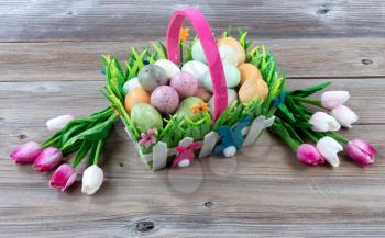 Carry basket filled with colorful eggs and pink tulips on weathered wooden boards for Easter background  
