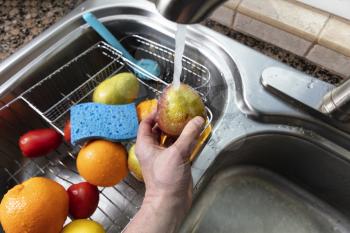 Male hand holding fresh pear fruit under running water from kitchen sink faucet. Clean or thoroughly washing concept 