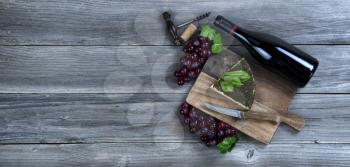 Fresh cheese wedge with a bottle of red wine plus basil leaves and grapes on rustic wood with copy space available 