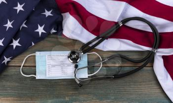 American flag with covid 19 Vaccination record card, personal facemask and stethoscope on fade blue rustic wooden planks 