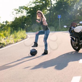 Young beautiful blonde girl in trendy jeans and a black T-shirt took off and put his helmet on the road near the modern motorcycle. Outdoor portrait in soft sunlight.
