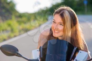 Young beautiful blonde girl in black t-shirt is resting on nature in seat of modern motorcycle. Outdoor portrait in soft sunny tones.