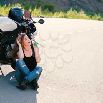 Young beautiful blonde girl in trendy jeans and a black t-shirt sits and drinks water near the modern motorcycle. Outdoor portrait in soft sunlight.