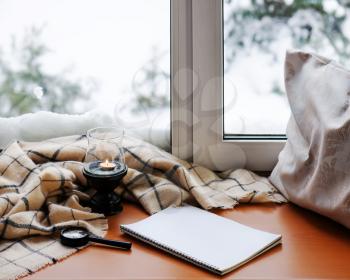Open notepad, magnifier glass, pillow, candle and beige warm plaid located on stylized wooden windowsill. Winter concept of comfort and relaxation.