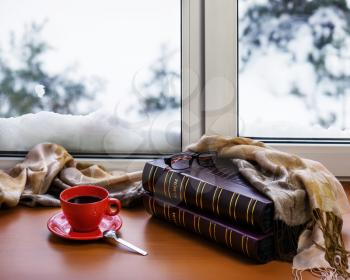Red cup of coffee or tea with a metal spoon, photo albums and glasses located on a stylized wooden windowsill. Winter concept of comfort and relaxation.