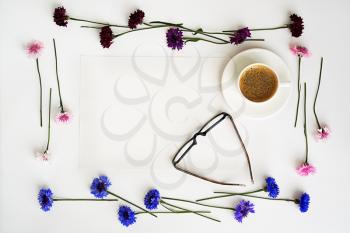 Paper, cup of coffee and glasses with wreath frame from petals of wildflowers on white background. Overhead view. Flat lay.