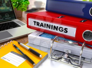 Red Ring Binder with Inscription Trainings on Background of Working Table with Office Supplies, Laptop, Reports. Toned Illustration. Business Concept on Blurred Background.