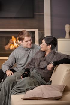 Young couple hugging on sofa in front of fireplace at home, looking at each other, smiling.